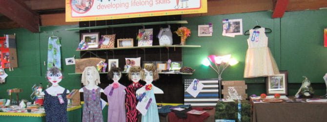 wide shot of 4-h fair projects including sewn garments on mannequins, art, photography, terrarium, wooden police flag under a banner that states 4-H is developing lifelong skills.